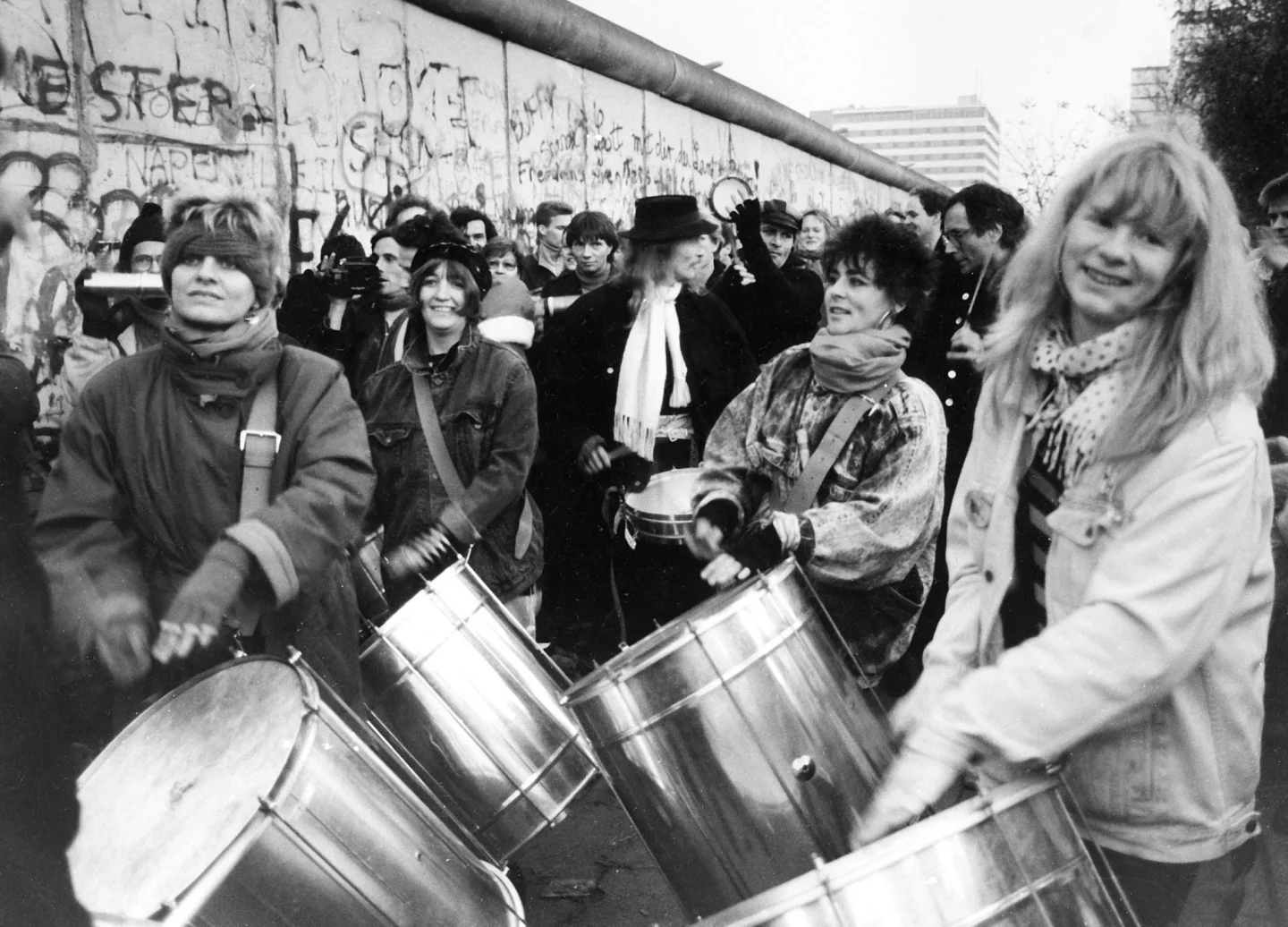 Schwarz-Weiß-Aufnahme zeigt fröhliche Trommlerinnengruppe, im Hintergrund viele Menschen udn die Berliner Mauer