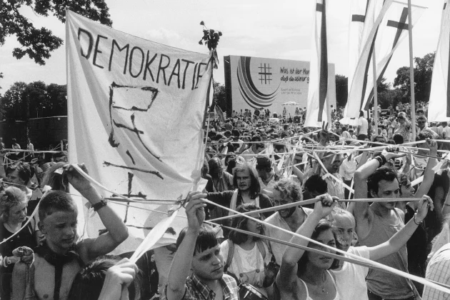 historisches Foto einer Demonstration in Leipzig, viele junge Menschen tragen mit erhobenen Armen Bänder, die sie zusammenhalten und Protestbanner mit Schlagwort Demokraite und christlichen Kreuzen