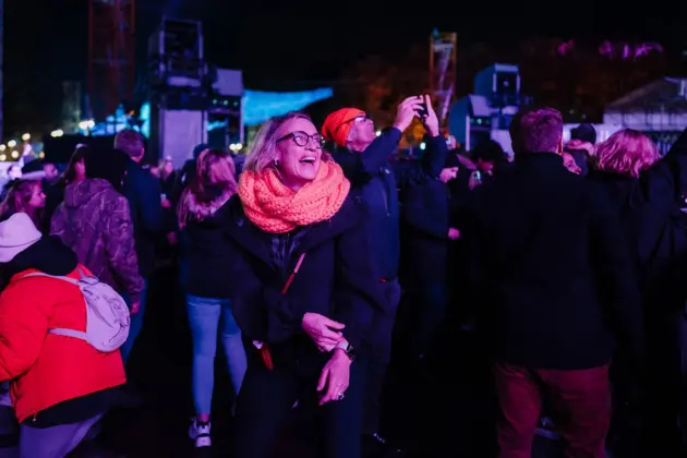 Fröhlich draußen feiernde Menschen, im Vordergrund lachende Frau mit orangeem Schal und älterer Mann macht Fotos auf dem Handy