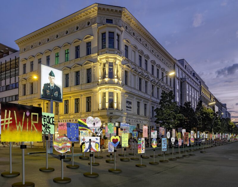 Visualisierung der Plakat-Installation im Berliner Stadtraum. Viele bunte Protestschilder stehen auf runden Füßen und Ständern in einer Reihe vor dem Checkpoint Charlie in der Dämmerung.