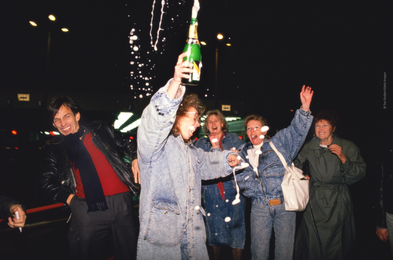 Eine Frau verspritzt in der Nacht jubelnd Sekt aus einer Flasche, 3 weitere Frauen stehen lachend hinter ihr, ein Mann dreht sich lachend weg