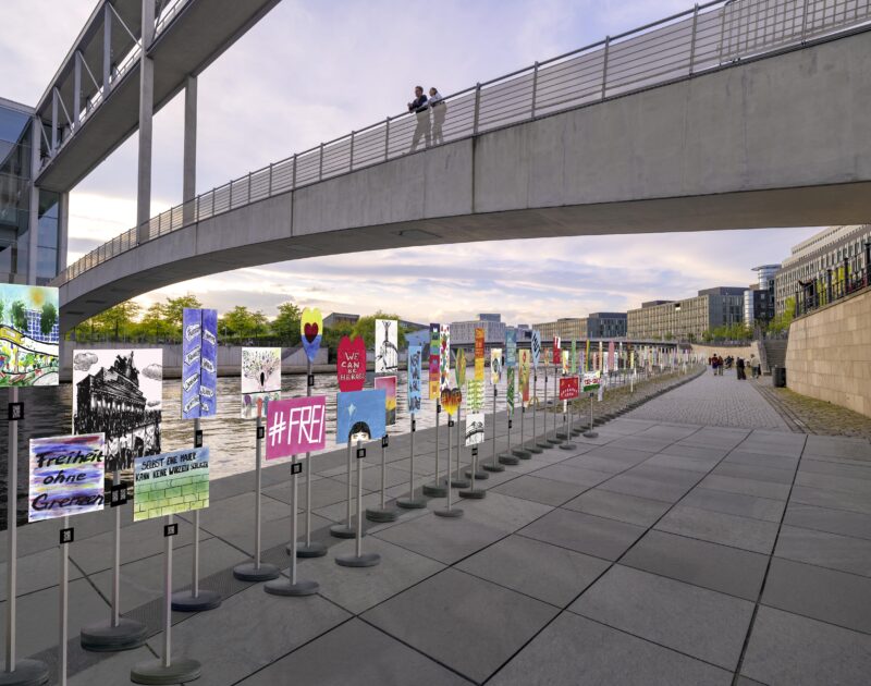 Visualisierung der Plakat-Installation im Berliner Stadtraum. Viele bunte Protestschilder stehen auf runden Füßen und Ständern in einer Reihe am Kapelle-Ufer.