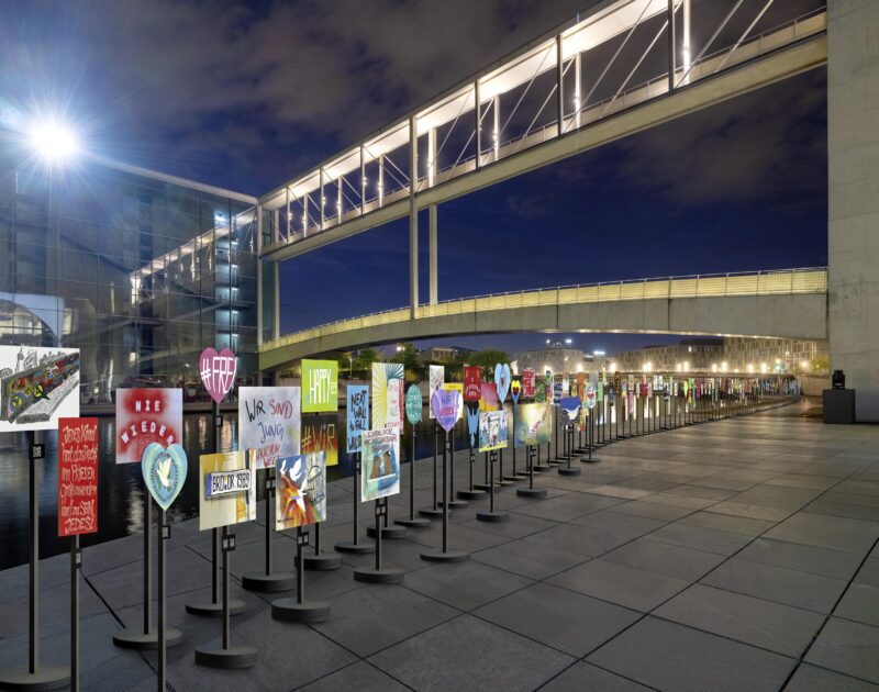 Visualisierung der Plakat-Installation im Berliner Stadtraum. Viele bunte Protestschilder stehen auf runden Füßen und Ständern in einer Reihe am Kapelle-Ufer bei Nacht.