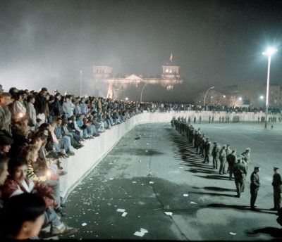 Menschenmassen sitzen auf der Berliner Mauer und schauen auf einen leeren Platz, auf dem Uniformierte eng nebeneinander stehen. Im Hintergrund sieht man den Bundestag.