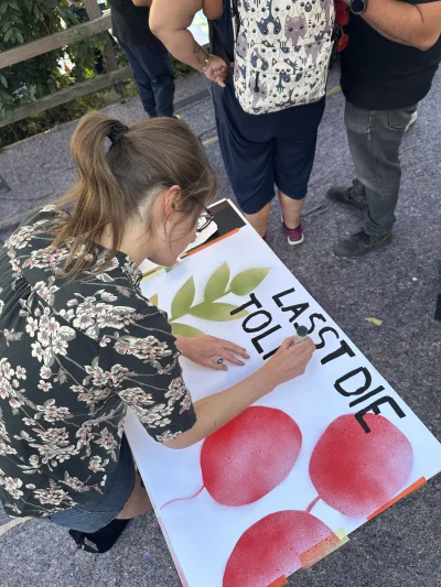 Junge Frau in sommerlichen Blumenshirt gestaltet Plakat mit roten Luftballons und Pflanze, darüber schreibt sie gerade "Lasst die Tol"