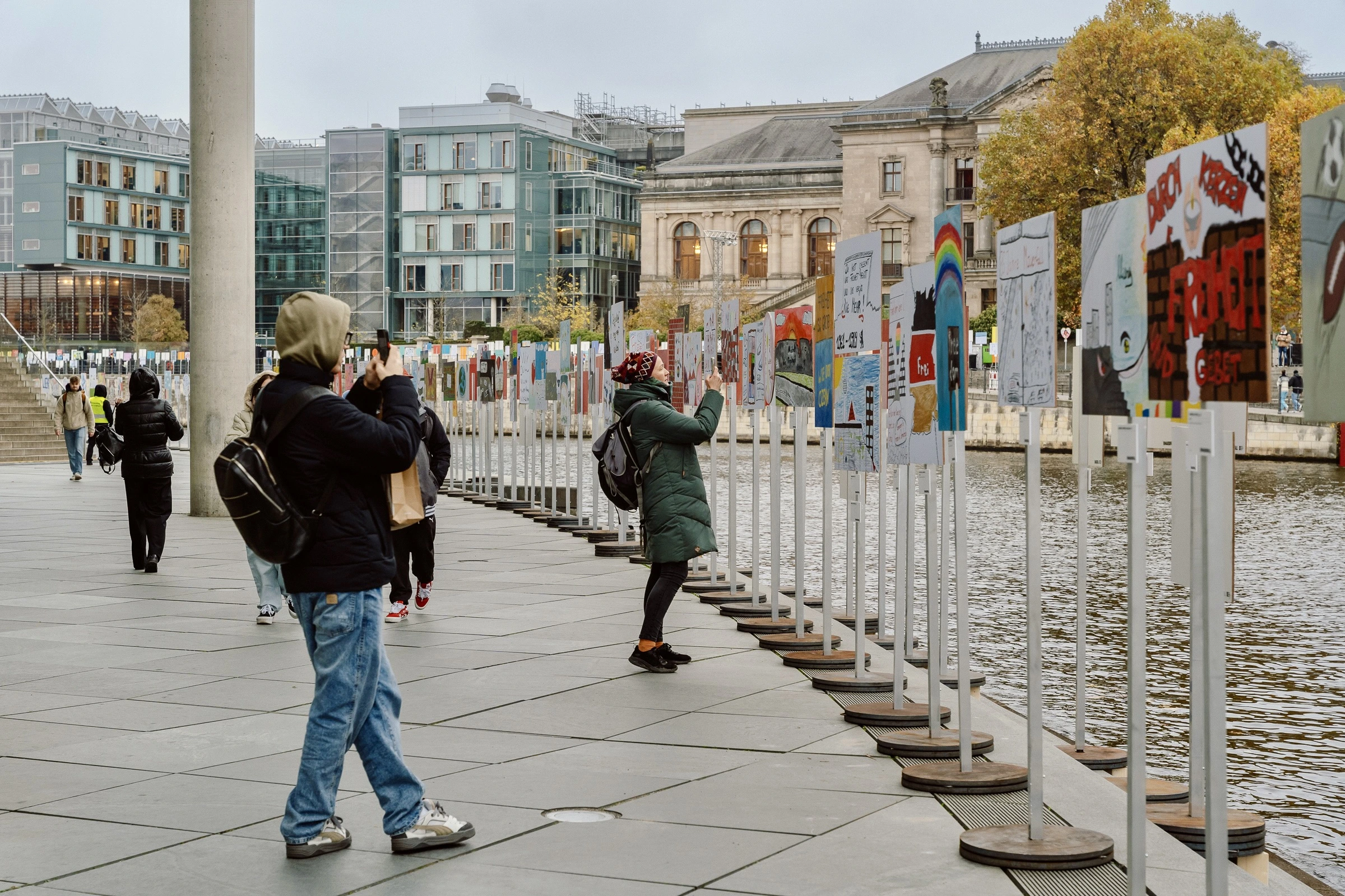 People standing before a colorful set of posters by the river and taking fotos